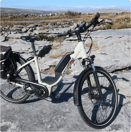 An e-Whizz Electric bike with a white frame and black accents is parked on a rocky, sunlit landscape, surrounded by grass and low stone walls. A small black bag is attached to the rear of the bike.