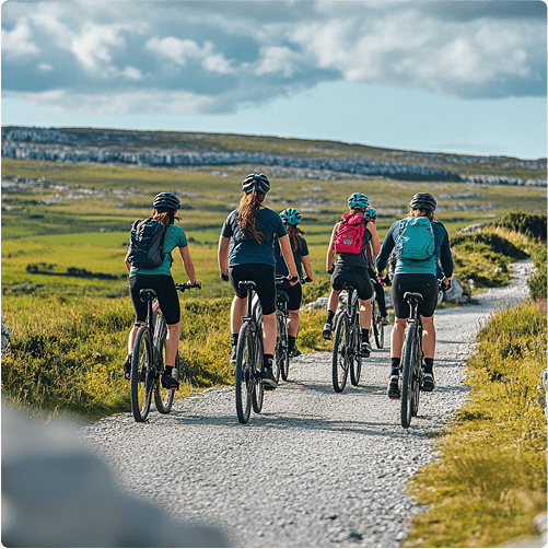 Five cyclists wearing helmets and backpacks ride e-Whizz Electric bikes along a gravel path through a scenic, green landscape under a partly cloudy sky. The group is heading away from the camera, enjoying an outdoor adventure.