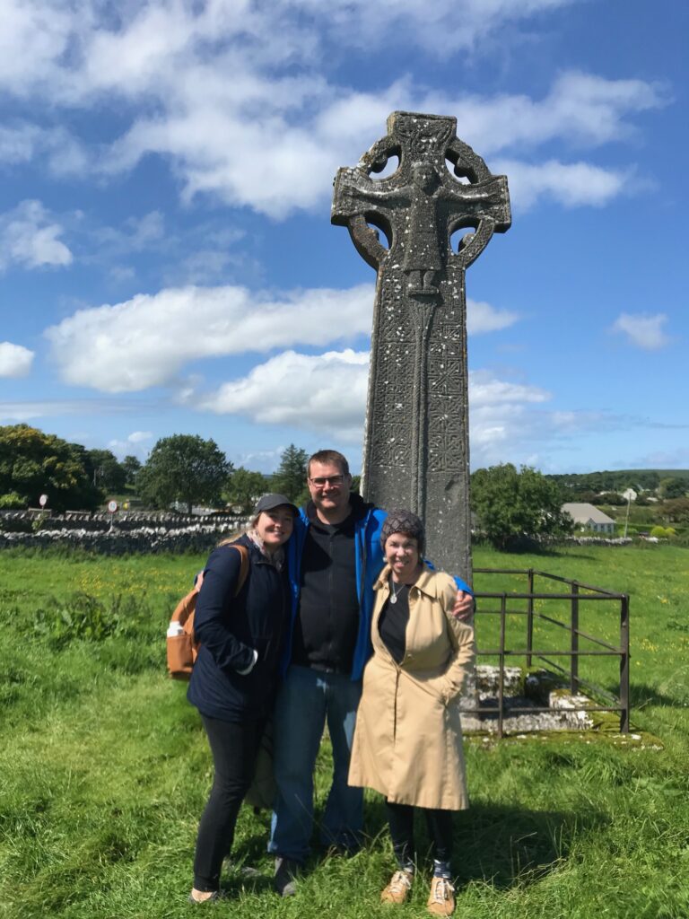 Three people stand together smiling in front of a large, ancient Celtic cross in a grassy field under a blue sky with scattered clouds. Trees, a stone wall, and distant buildings are visible in the background.