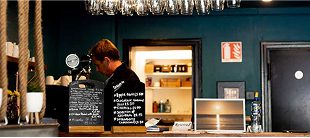 A man stands behind a counter in a cozy cafe or bar, with glasses hanging overhead, a chalkboard menu, and a laptop displaying a sunset image—perhaps planning his next ride on an e-Whizz Electric bike. Shelves and a doorway are in the background.