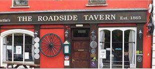 A red-painted pub exterior with the sign The Roadside Tavern Est. 1865, a large wagon wheel, several plaques, and white-framed windows—ideal for a stop when touring the area with e-Whizz Electric bikes.