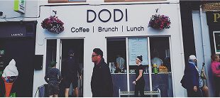 People walk past a white café with the sign “DODI Coffee | Brunch | Lunch.” Hanging flower baskets adorn the entrance, while several people and e-Whizz Electric bikes are visible both inside and outside the shop.