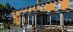 A large yellow two-story house with many windows, a sunroom, and potted plants on a paved entryway under a blue sky, perfect for parking your e-Whizz Electric bikes after an afternoon ride.