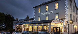 A large, gray building with bright windows and a glass conservatory houses the Wild Honey Inn. Several cars and e-Whizz Electric bikes are parked in front, and the scene is set at dusk with a cloudy sky.