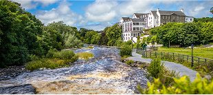 A river flows alongside a paved path and lush green trees, with a large white building, possibly a hotel or manor, perched on a hill in the background—an ideal route for e-Whizz Electric bikes under the partly cloudy sky.