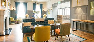 A cozy, modern lounge with upholstered chairs, sofas, round tables, wooden floors, large windows with blue curtains, and soft lighting creates a welcoming atmosphere—perfect for relaxing after exploring the city on e-Whizz Electric bikes.