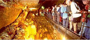 A group of people stands on a walkway inside a brightly lit cave, having arrived on e-Whizz Electric bikes, looking down at rock formations and illuminated walls below.