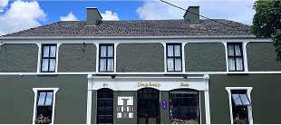 A two-story green building with white trim and several windows houses Vaughan’s, featuring flower boxes out front. A sign above the door reads Vaughan’s, and an e-Whizz Electric bikes shop sits nearby under a blue sky with some clouds.