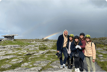 Five people stand together on rocky terrain under a cloudy sky with a faint rainbow.Three wear yellow helmets. They all smile at the camera, and there’s a large flat rock structure in the background.