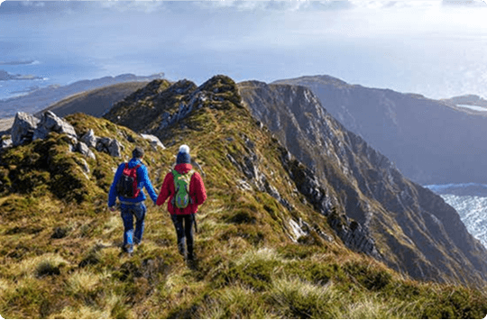 Two hikers wearing backpacks and outdoor gear walk along a narrow, grassy mountain ridge with dramatic cliffs, overlooking the ocean and distant islands under a cloudy sky—the ideal landscape to explore with e-Whizz Electric bikes.