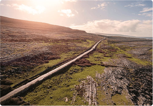 A narrow road winds through a rocky, barren landscape covered with patches of green grass under a partly cloudy sky, with sunlight illuminating the scene from the left—perfect for exploring on e-Whizz Electric bikes.