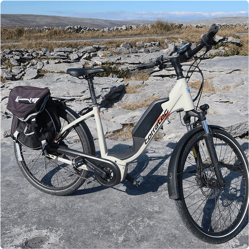 A light-colored e-Whizz Electric bike with a black saddlebag is parked on a rocky surface, with dry grass and distant hills visible in the background under a clear sky.