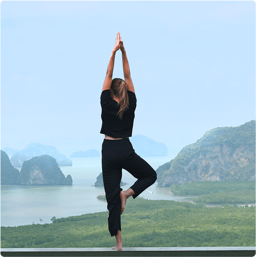 A woman practices yoga in a tree pose on a platform, overlooking lush green hills and distant mountains under a clear sky—an ideal spot to explore with e-Whizz Electric bikes before unwinding with some mindful stretching.