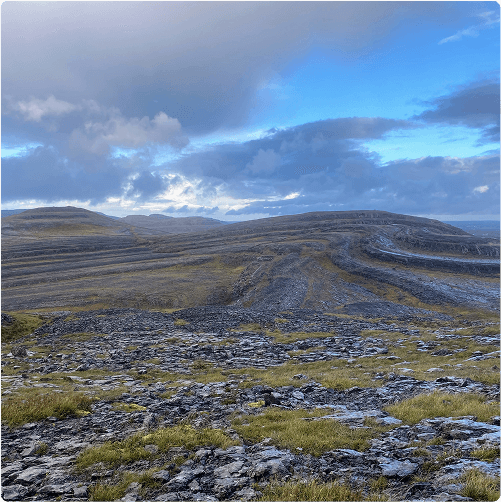 Rocky, undulating landscape with sparse grass, leading to distant hills under a partly cloudy sky—perfect for exploring by e-Whizz Electric bikes. The remote, windswept scene invites adventure with sunlight breaking through the clouds.