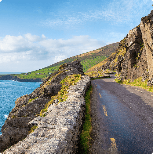 A narrow coastal road winds along rocky cliffs overlooking the blue ocean, with green hills and a partly cloudy sky in the background.
