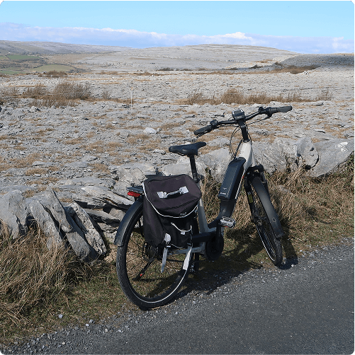 A black e-Whizz Electric bike with a saddlebag rests against a stone wall beside a rural road, with rocky terrain and rolling hills stretching out under a blue sky in the background.