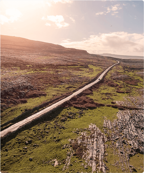 A narrow road winds through a rocky, green landscape under a bright sky, perfect for exploring on e-Whizz Electric bikes, with sunlight illuminating the hills and scattered clouds in the distance.
