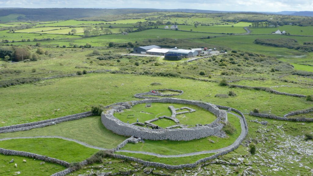 Aerial view of a large stone ring fort surrounded by green fields, with smaller stone walls and farm buildings in the background under a partly cloudy sky.