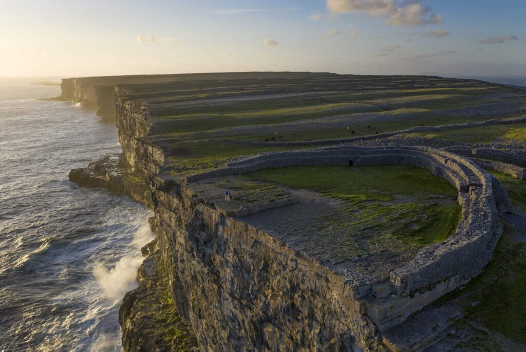 Aerial view of a circular stone fort on a high sea cliff, overlooking the ocean at sunset, with waves crashing below and green fields stretching into the distance.