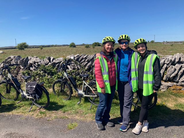 Three people wearing helmets and green safety vests stand smiling together by a stone wall, with bicycles parked nearby on a sunny day. Theres open countryside and a clear blue sky in the background.