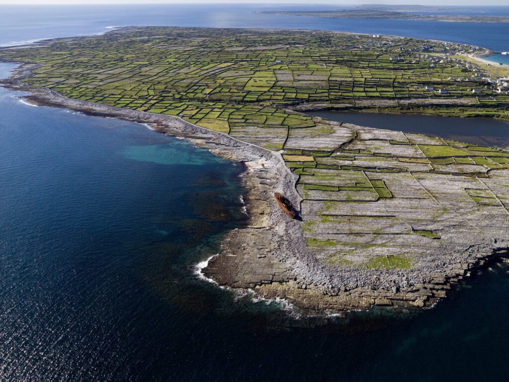 Aerial view of a rocky coastline with patchwork green fields divided by stone walls, surrounded by blue ocean, and a rusted shipwreck near the shore. Scattered buildings are visible in the distance.