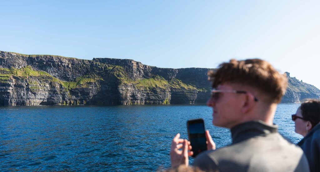 A local guide stands on a boat with a traveler holding a smartphone, both set against steep cliffs and clear blue water under a bright sky.