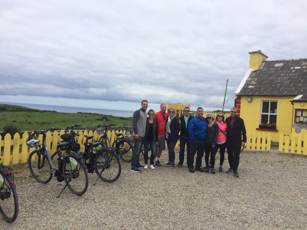 A group of eight people stand smiling beside bicycles in front of a yellow cottage with a picket fence. A grassy landscape and the sea are visible under a cloudy sky in the background.